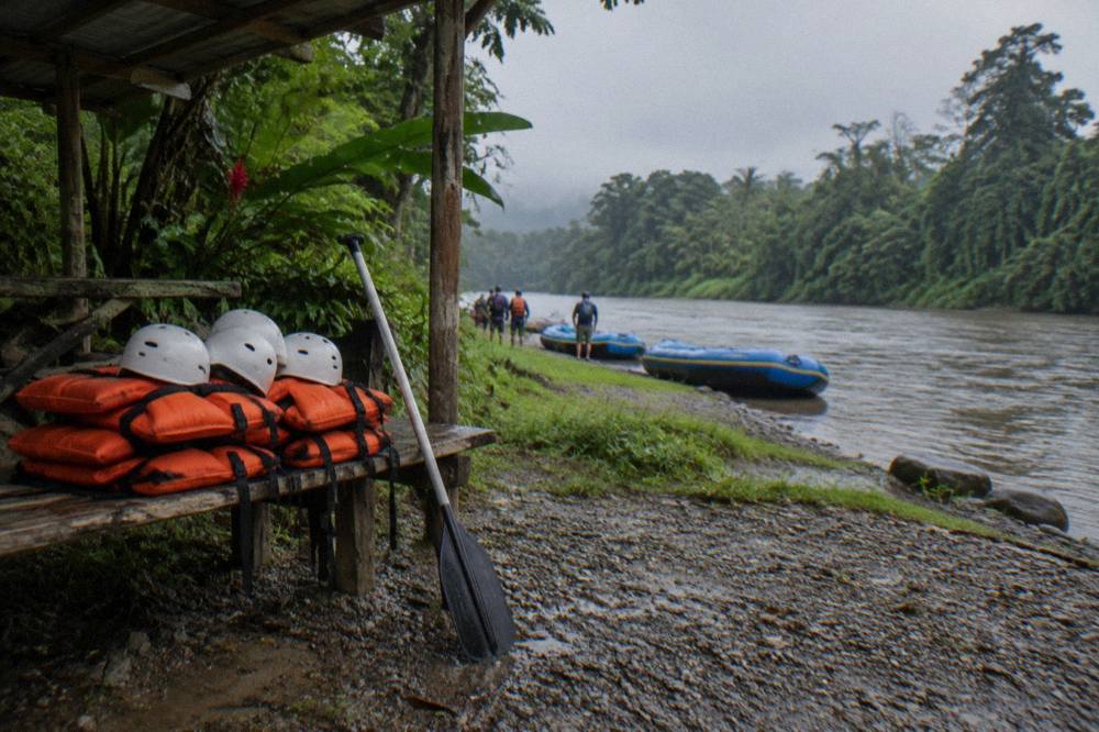 A stack of orange life jackets and white helmets on a wooden bench at a river put-in