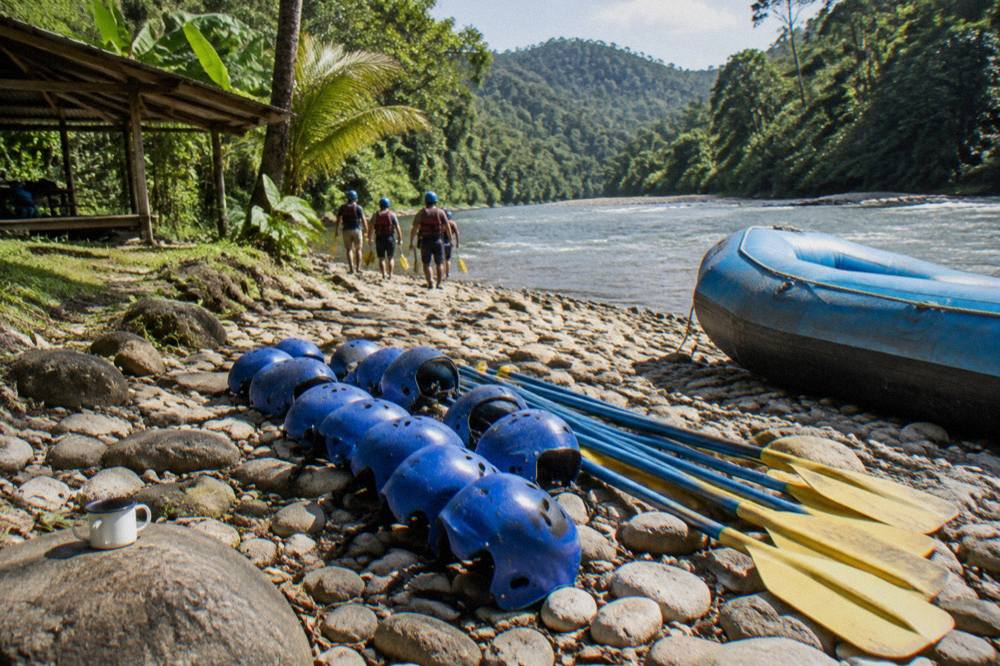 Rafting helmets and paddles laid out on a riverbank beside an inflated raft
