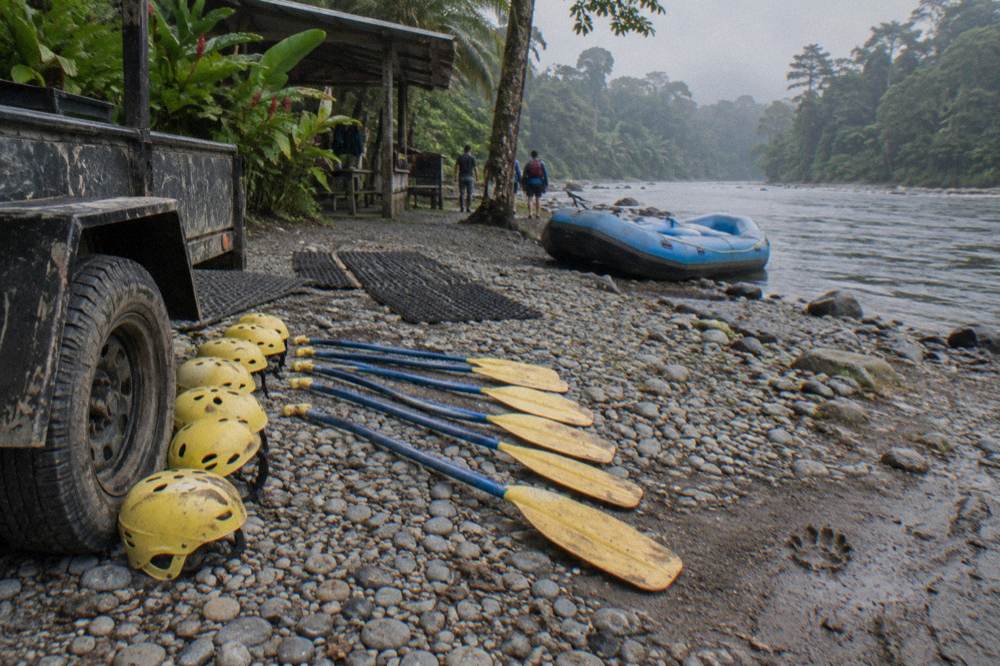 Rafting helmets and paddles laid out on a riverbank beside a trailer