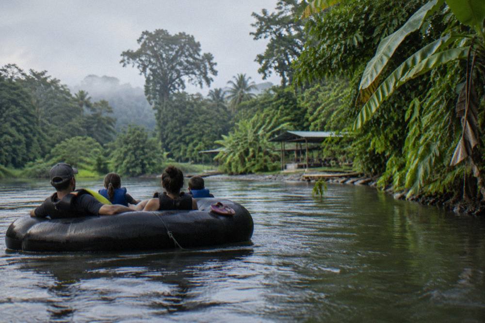 A raft with paddlers in helmets and life jackets floating past tall rainforest trees along a calm river stretch
