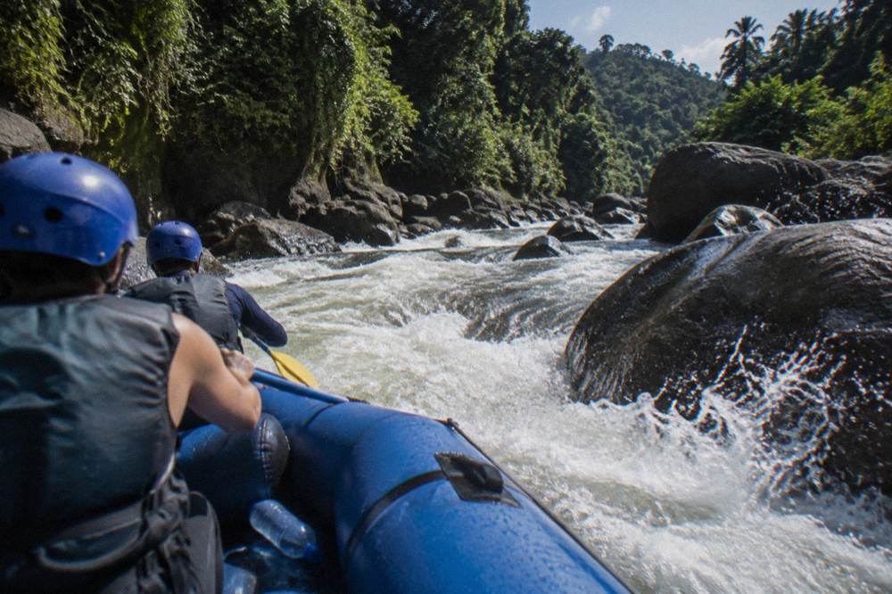 A guided raft passing through a churning rapid between boulders in a green canyon