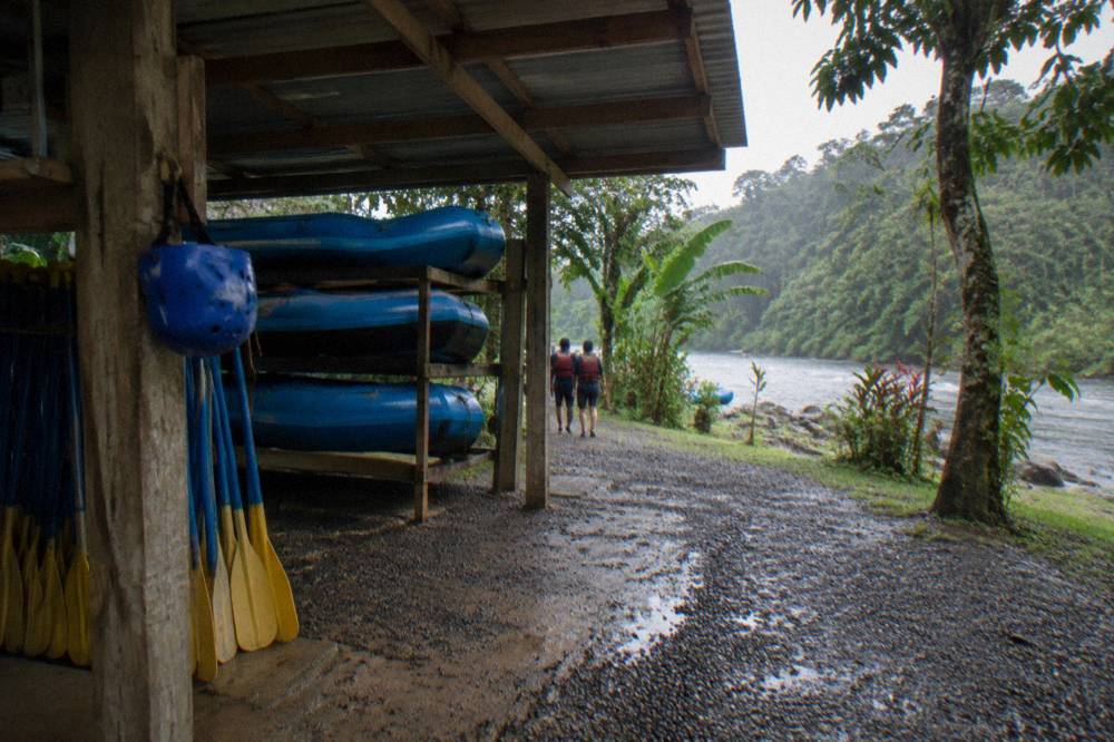 A covered open-air meeting area beside a river with rafts and paddles stacked ready
