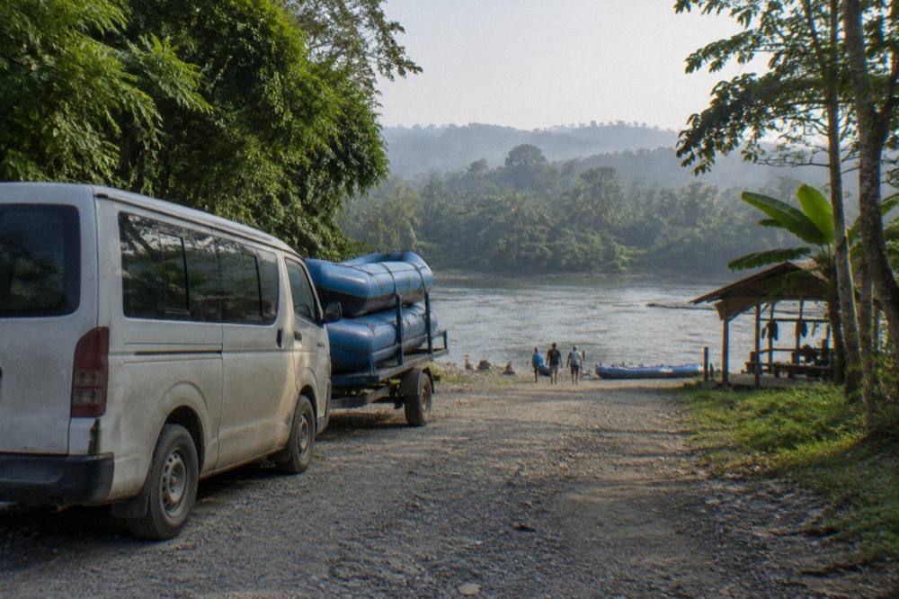 A van parked on a gravel road beside a tropical river with rafts loaded on a trailer