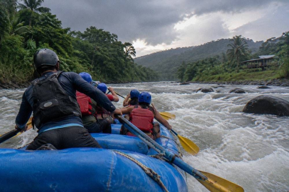 A guide steering from the rear of a raft as paddlers drive through churning whitewater