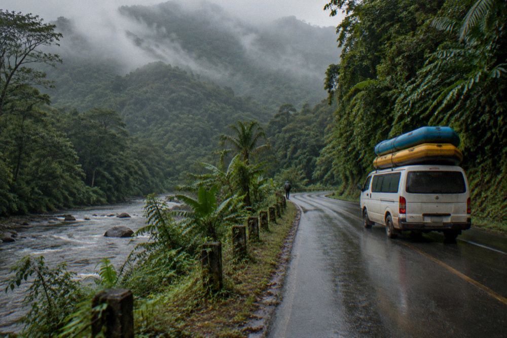A loaded rafting van on a winding road through rainy mountain forest