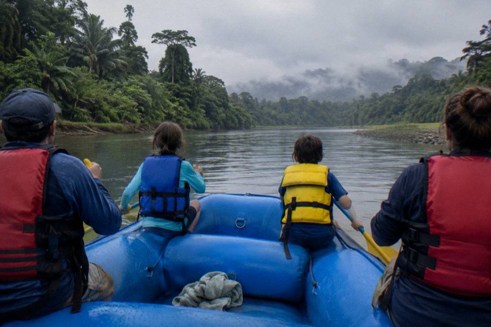 Family in life jackets paddling a raft on calm water between rainforest banks