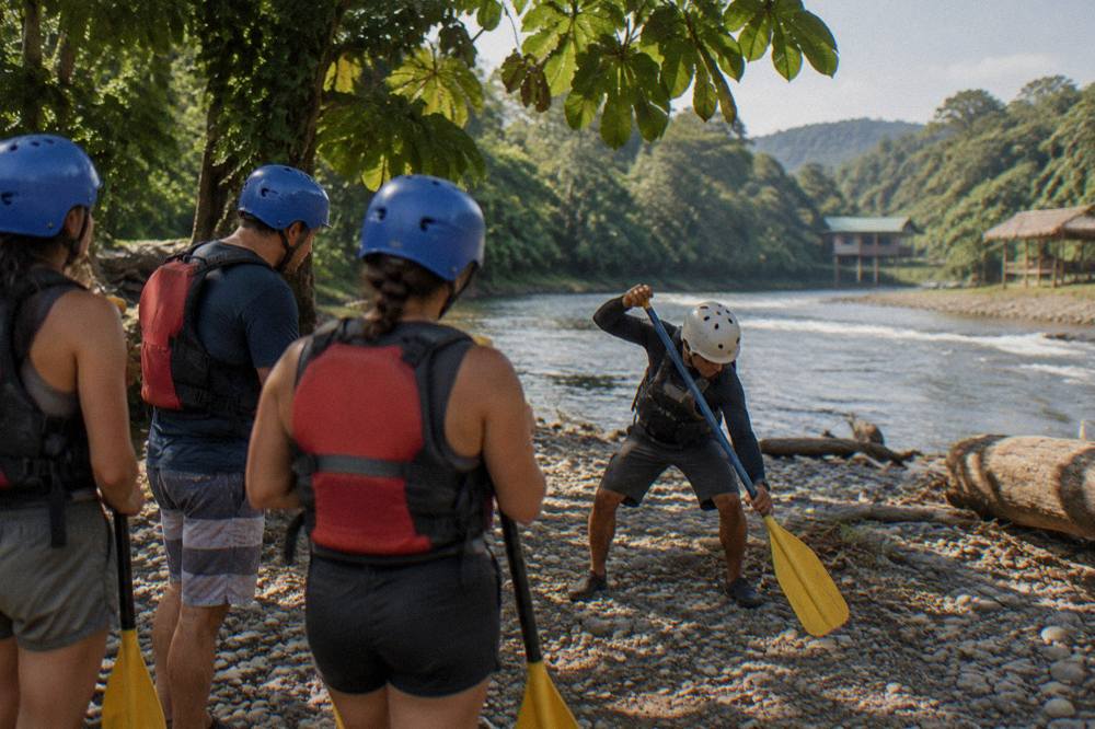 A guide demonstrating paddle technique to a small group wearing helmets and life jackets on a gravel riverbank
