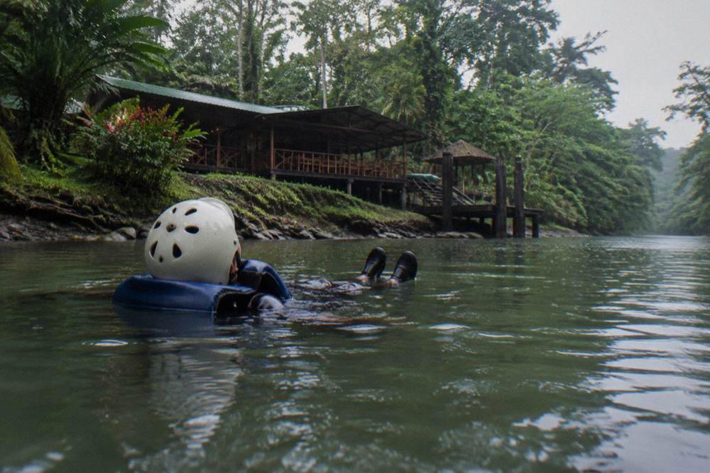A swimmer in a helmet and life jacket floating on their back with feet raised in a calm green river reach