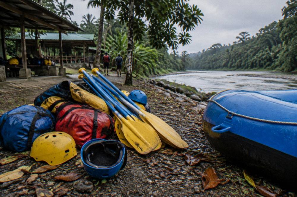 A pile of helmets, paddles and dry bags arranged on a riverbank beside a raft