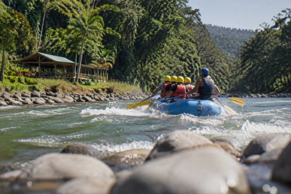 Raft with several paddlers in helmets and life jackets crossing a green jungle river rapid