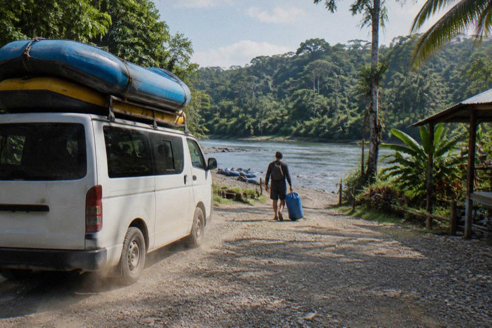 A rafting van parked beside a gravel river access road with rafts strapped on top