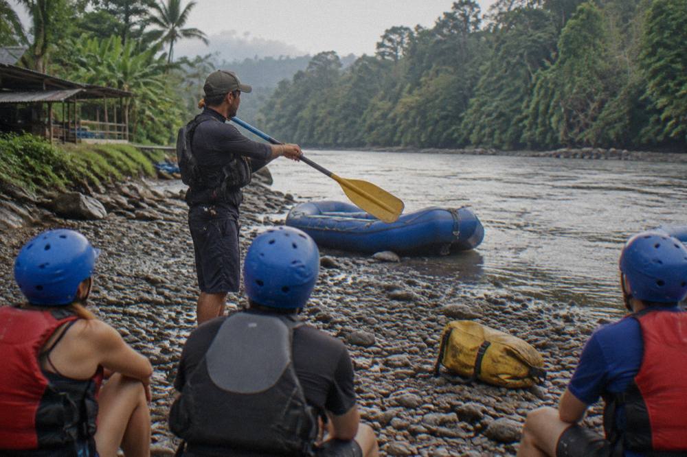 A certified rafting guide giving a paddle briefing to guests wearing helmets and vests on a riverbank
