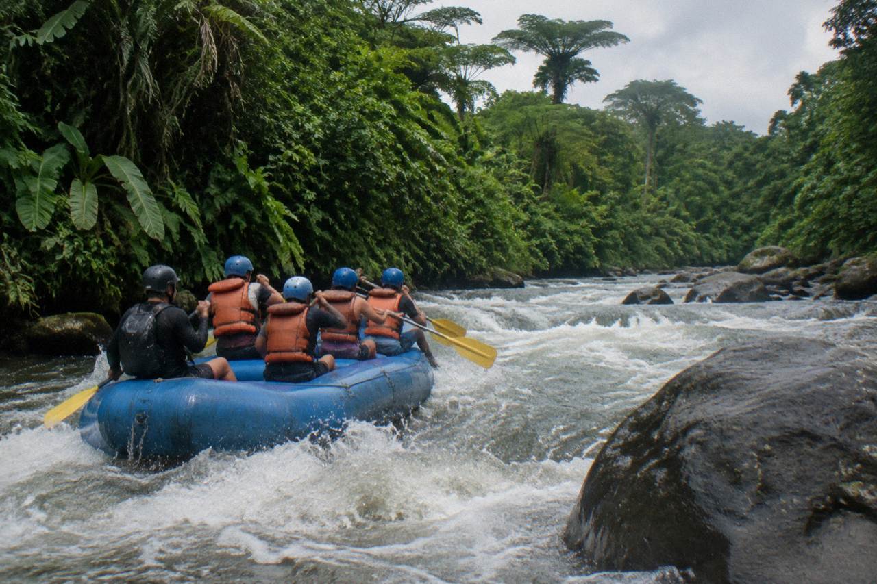 A rafting crew in helmets and life vests paddling a blue inflatable raft through a jungle rapids section
