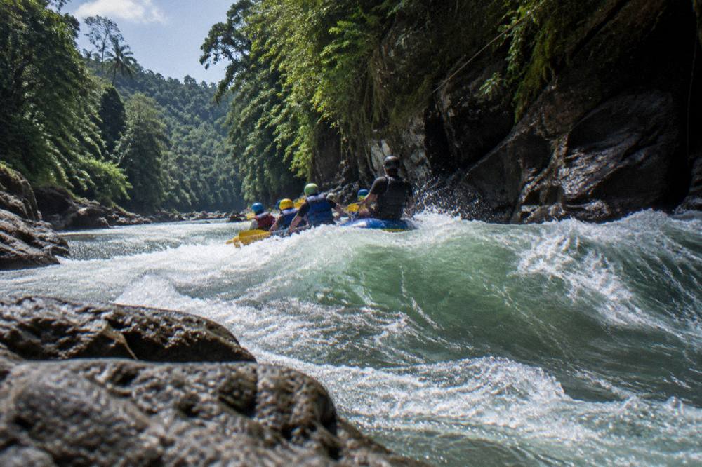 Raft cutting through a green wave in a jungle canyon with paddlers in helmets