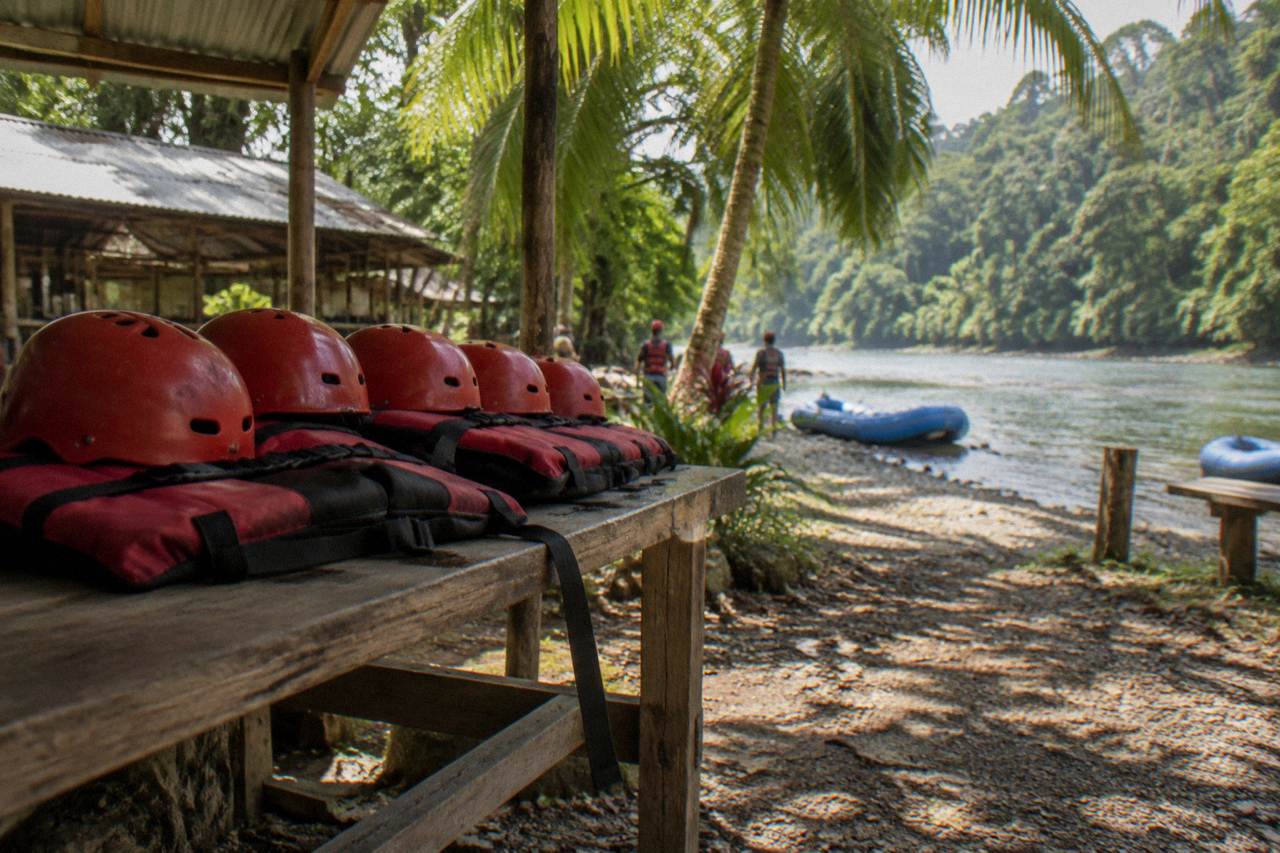 Rafting helmets and life jackets lined up on a riverbank gear table under jungle shade