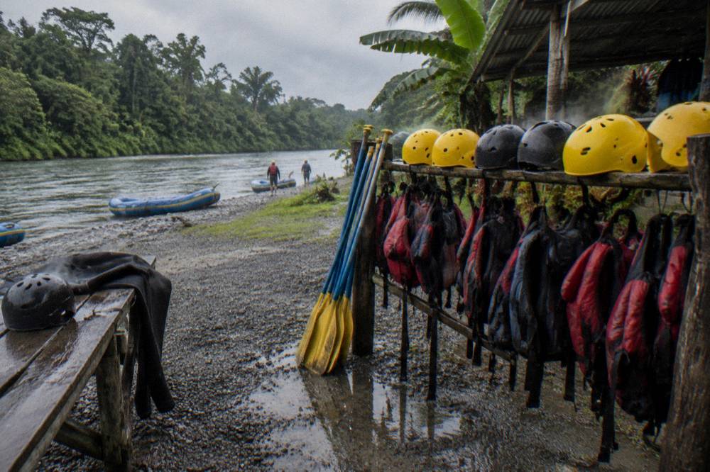 Wet rafting gear, helmets and paddles laid out on a riverbank under overcast light