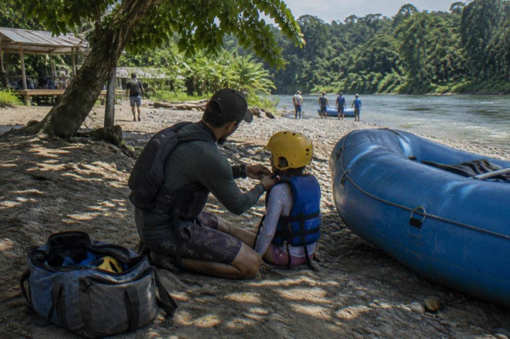 A guide fitting a helmet on a rafter beside a raft on a gravel riverbank
