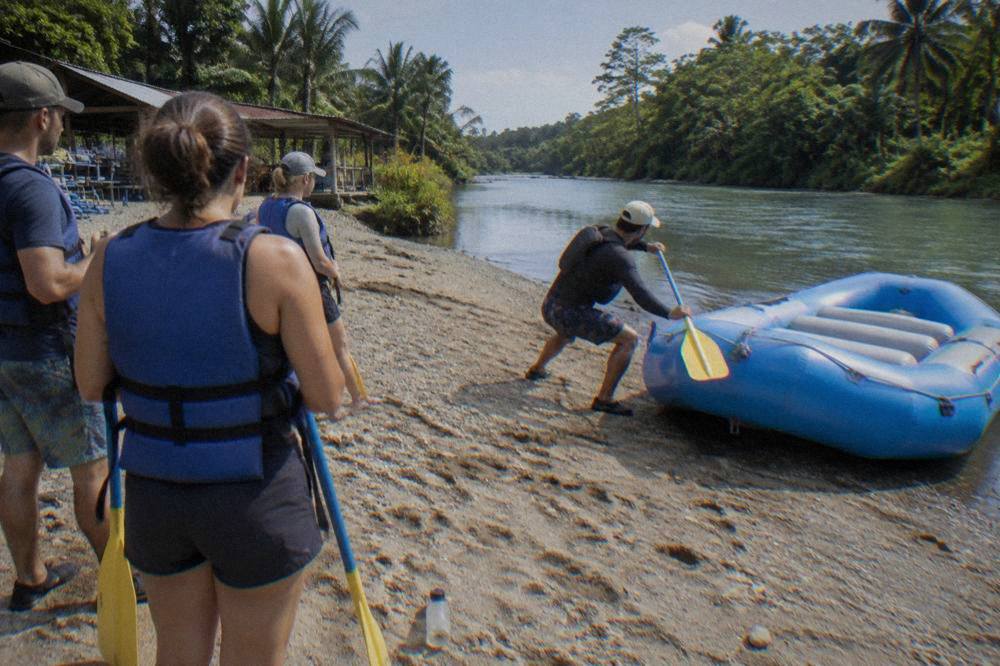 Raft guide giving a safety briefing to guests holding paddles beside a raft on a gravel river bank