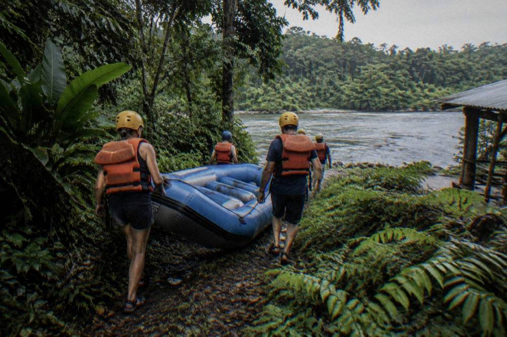 Guests in life jackets and helmets carrying a raft toward the water through green riverside vegetation