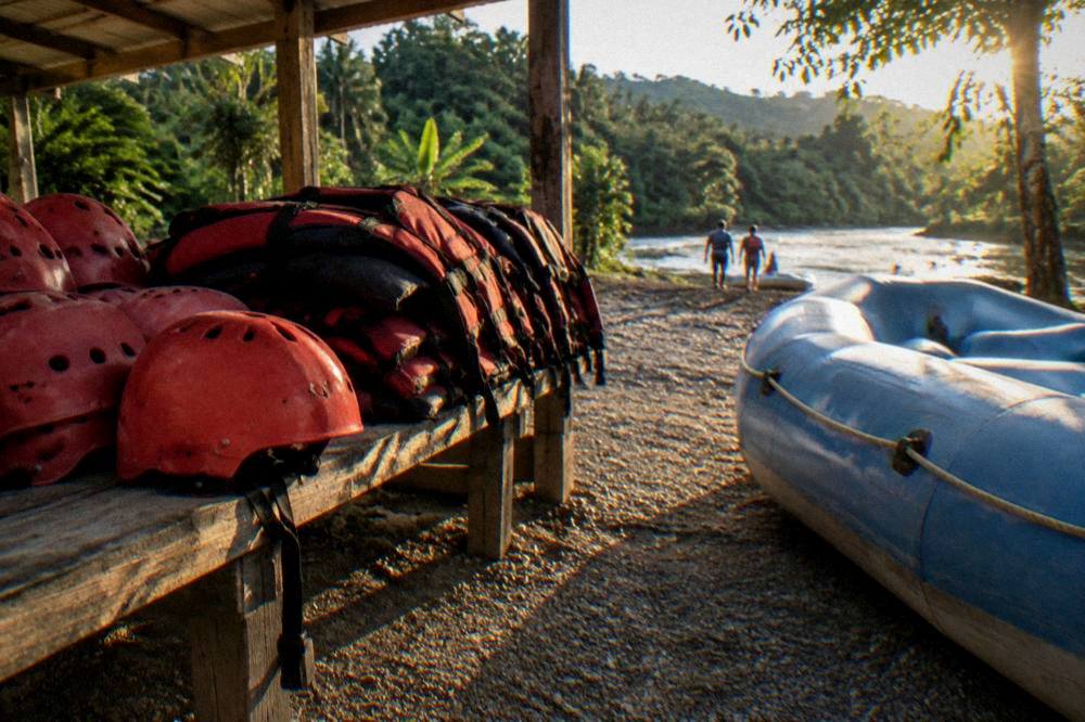 Stacked helmets and life jackets arranged on a riverside bench beside a raft