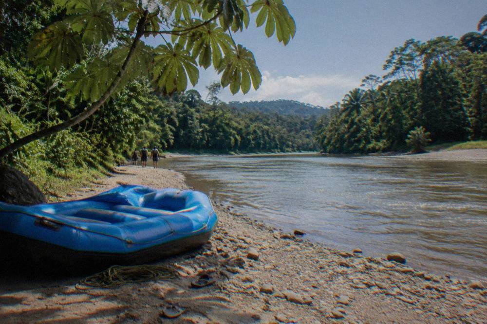 An inflatable raft resting on a gravel riverbank beside dense green rainforest