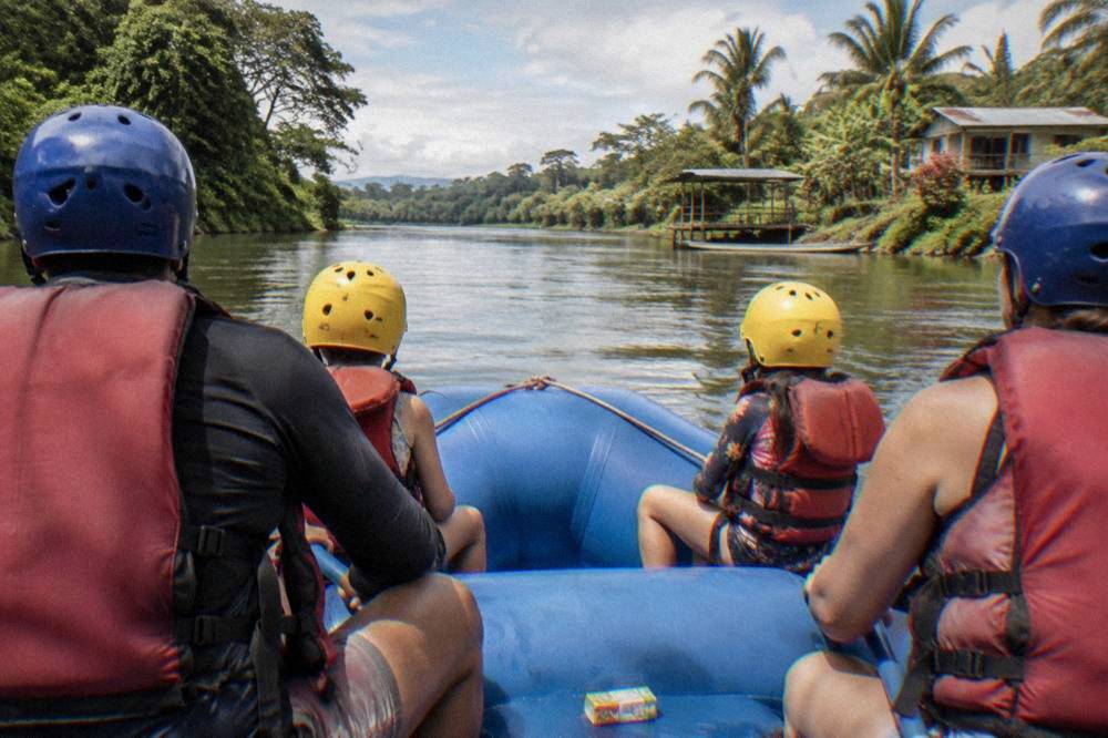 A family raft with paddlers in helmets and life jackets on flat water between green banks