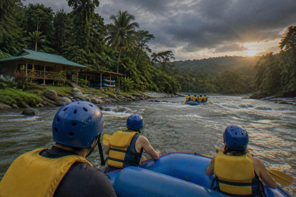 A raft with paddlers crossing a green tropical rapid between forested banks.