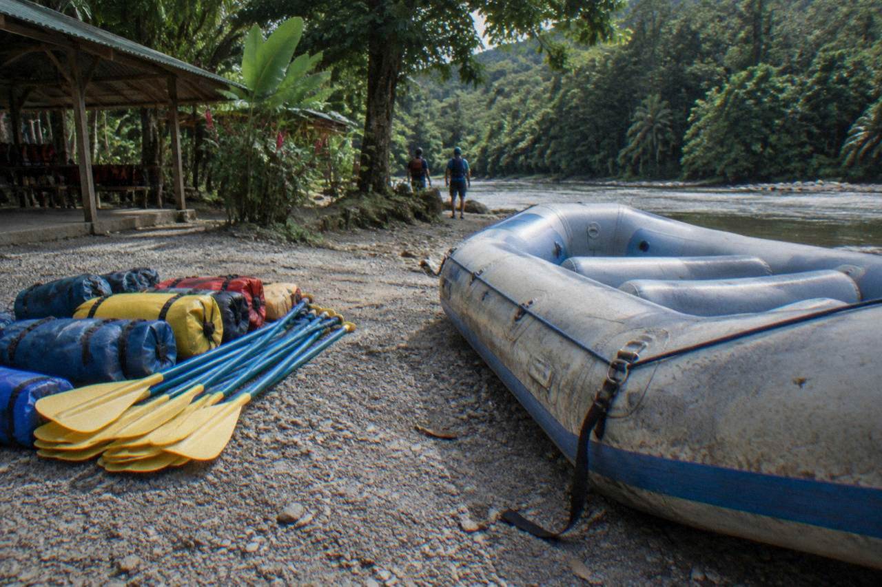 Dry bags and paddles stacked beside an inflatable raft on a gravel riverbank under jungle cover