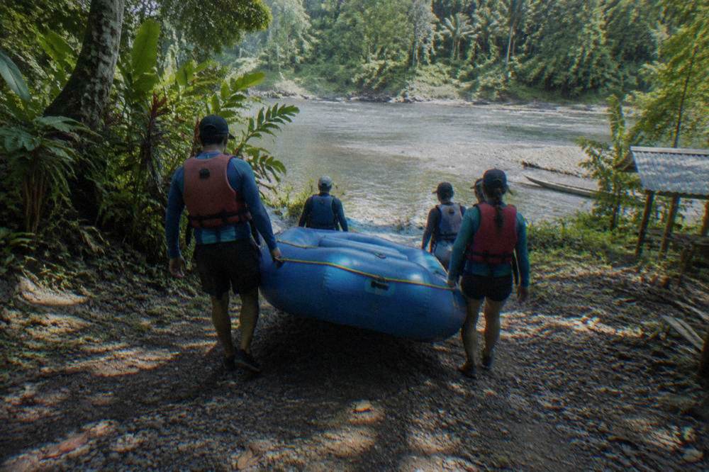Paddlers in rash guards and life jackets carrying a raft toward the water
