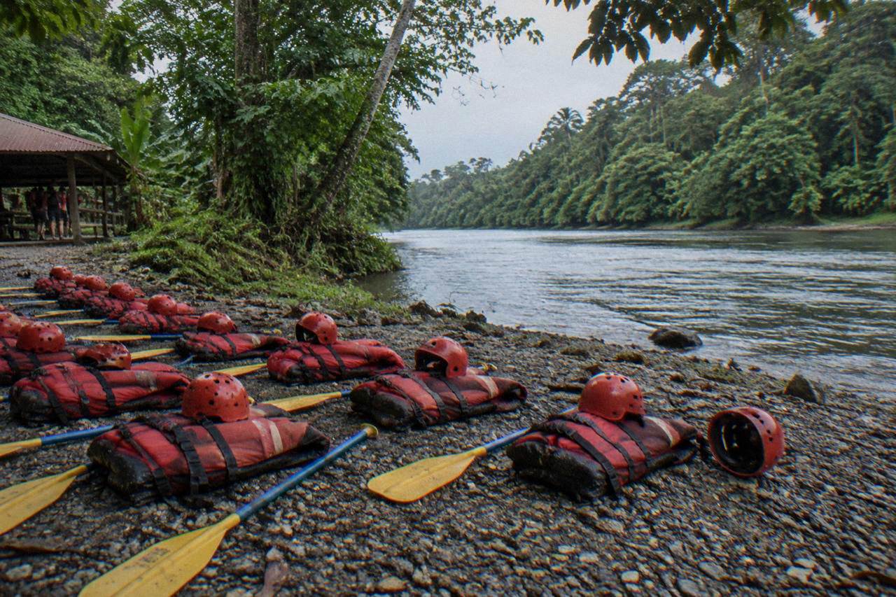 Rafting paddles, helmets and life jackets laid out on a riverbank under dense jungle canopy