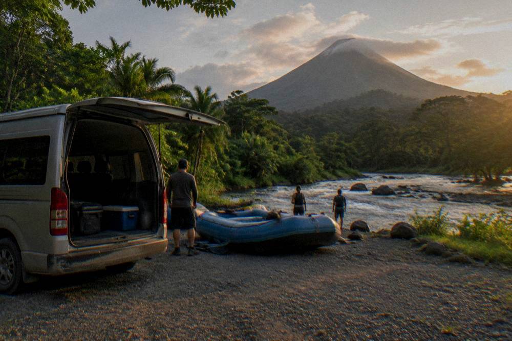 A van parked beside a river put-in area surrounded by rainforest