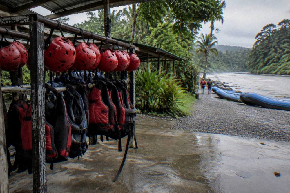 Stacked helmets and life jackets on a rack beside a river
