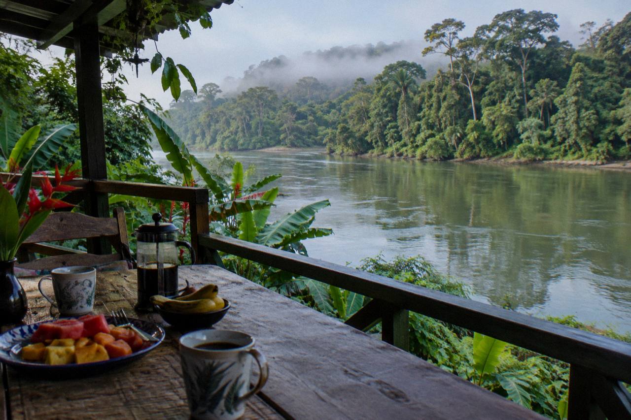 A waterside veranda with a table of fruit and coffee overlooking a calm green river and misty forest